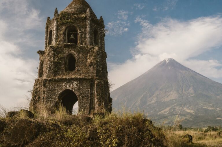 A weathered stone bell tower ruin with a volcano rising behind it
